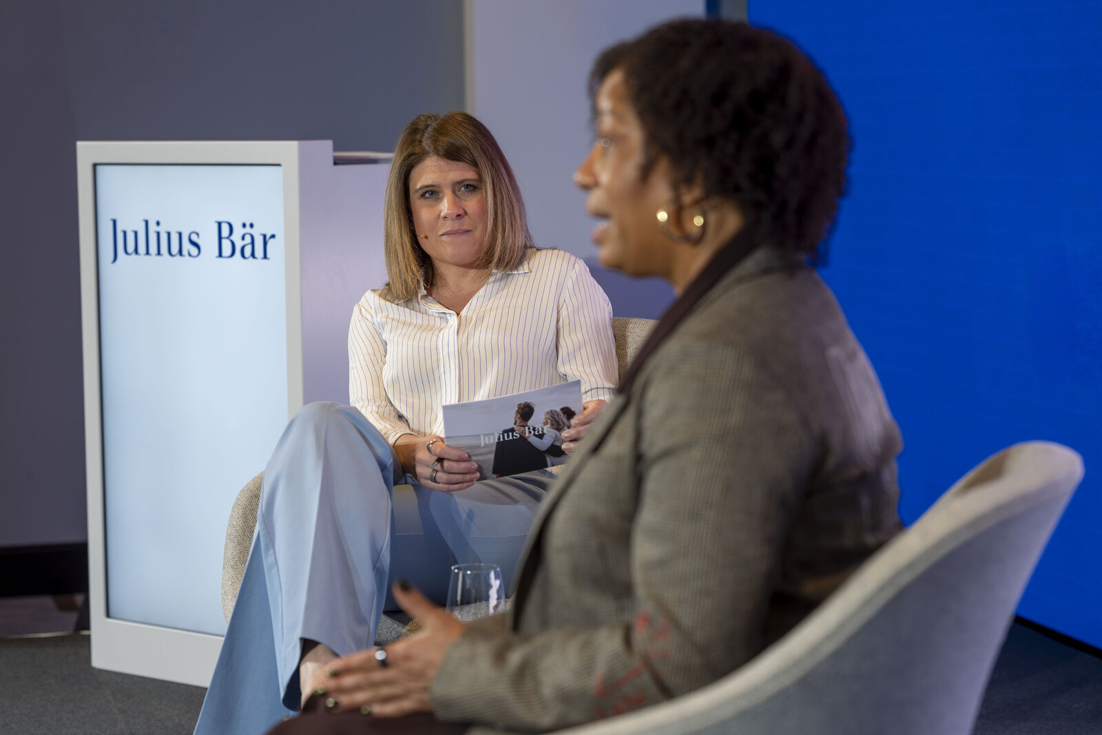 Two speakers in conversation on stage at a live event, one holding a printed programme