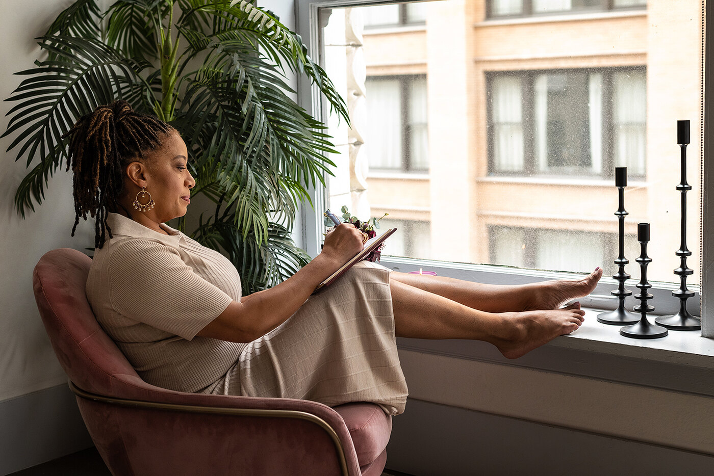 Lisane Basquiat sitting in a velvet armchair by a window, feet up on the sill, writing in a notebook