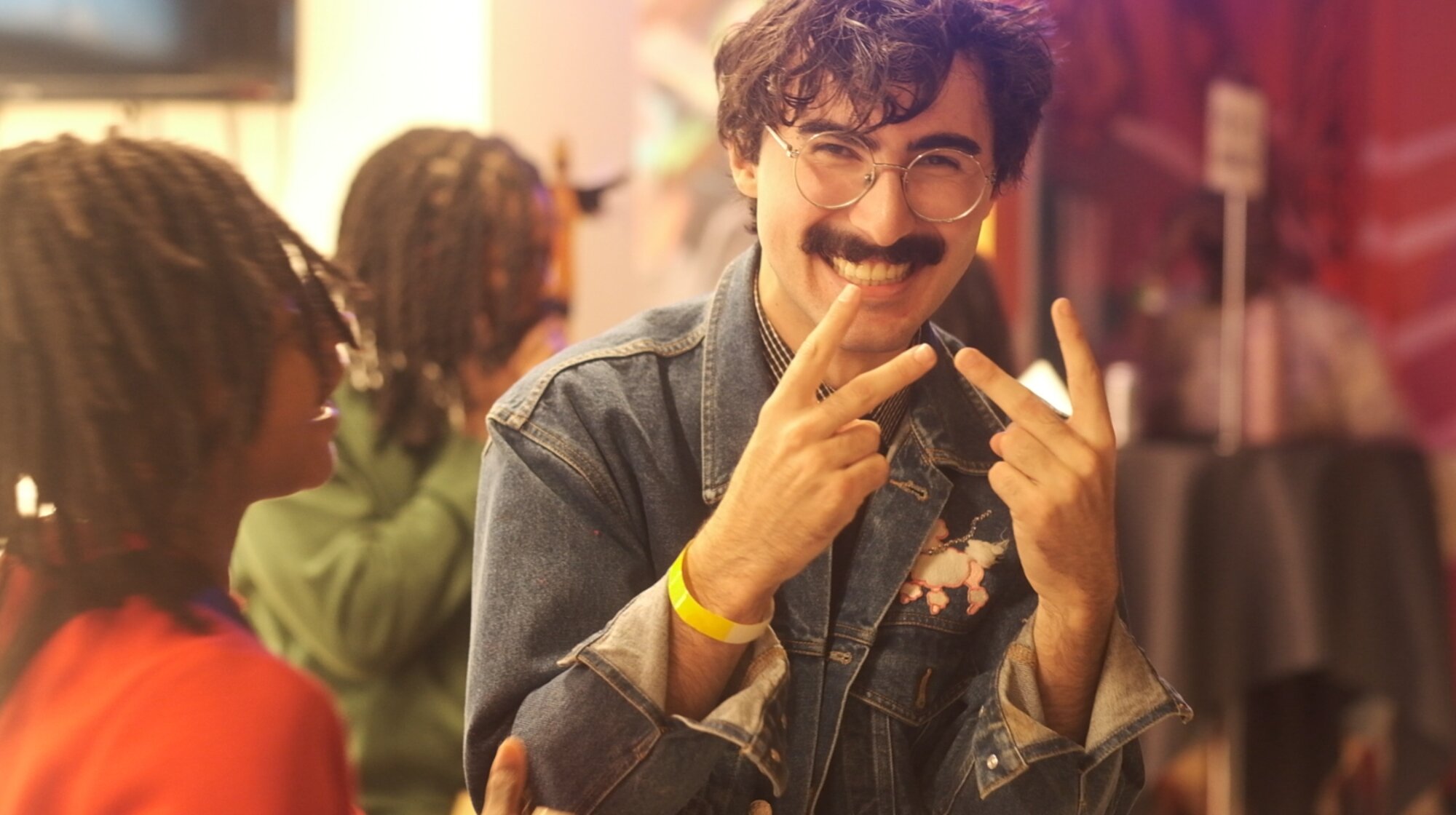 A guest smiling and throwing peace signs at a Soulful Saturdays gathering, others talking behind him